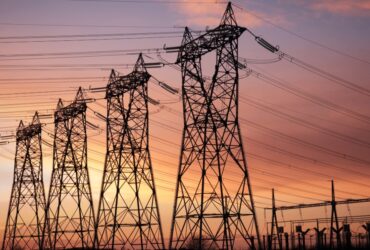 The sun is setting behind multiple high-voltage electrical transmission towers, with wires carrying electricity.