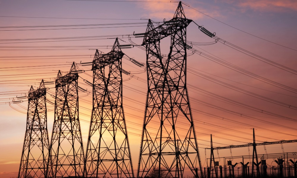 The sun is setting behind multiple high-voltage electrical transmission towers, with wires carrying electricity.