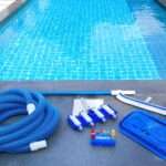 A close-up of a swimming pool with maintenance equipment lining one side. There's a brush, a net, tubing, and a water testing kit.