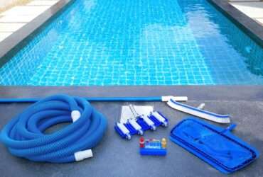 A close-up of a swimming pool with maintenance equipment lining one side. There's a brush, a net, tubing, and a water testing kit.