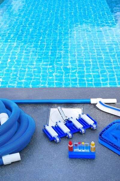 A close-up of a swimming pool with maintenance equipment lining one side. There's a brush, a net, tubing, and a water testing kit.
