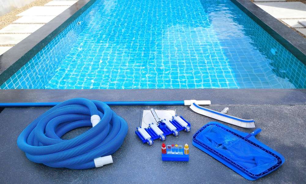 A close-up of a swimming pool with maintenance equipment lining one side. There's a brush, a net, tubing, and a water testing kit.
