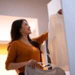 Woman smiles while hanging a white shirt in a closet, holding another top on a hanger in a warmly lit room.