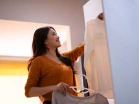 Woman smiles while hanging a white shirt in a closet, holding another top on a hanger in a warmly lit room.