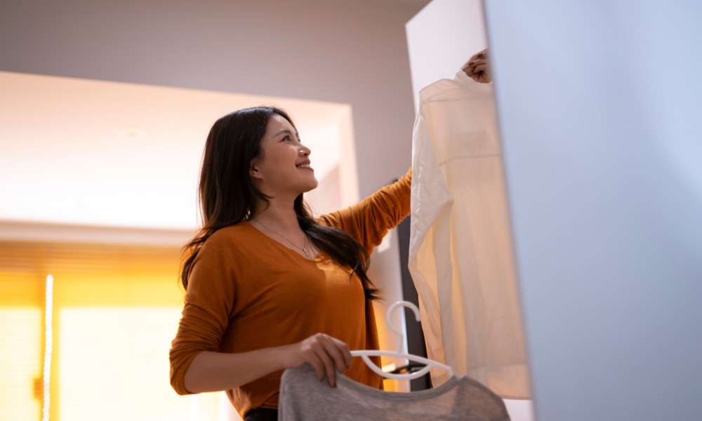 Woman smiles while hanging a white shirt in a closet, holding another top on a hanger in a warmly lit room.