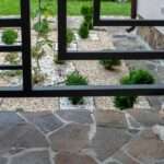 Fluffy white dog rests near a modern black fence overlooking a neat garden of gravel, stones, and shrubs.