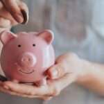 A close-up view shows a person placing a small copper coin into the slit on a small, pink piggy bank.