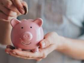 A close-up view shows a person placing a small copper coin into the slit on a small, pink piggy bank.