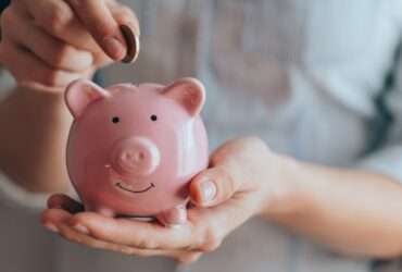 A close-up view shows a person placing a small copper coin into the slit on a small, pink piggy bank.
