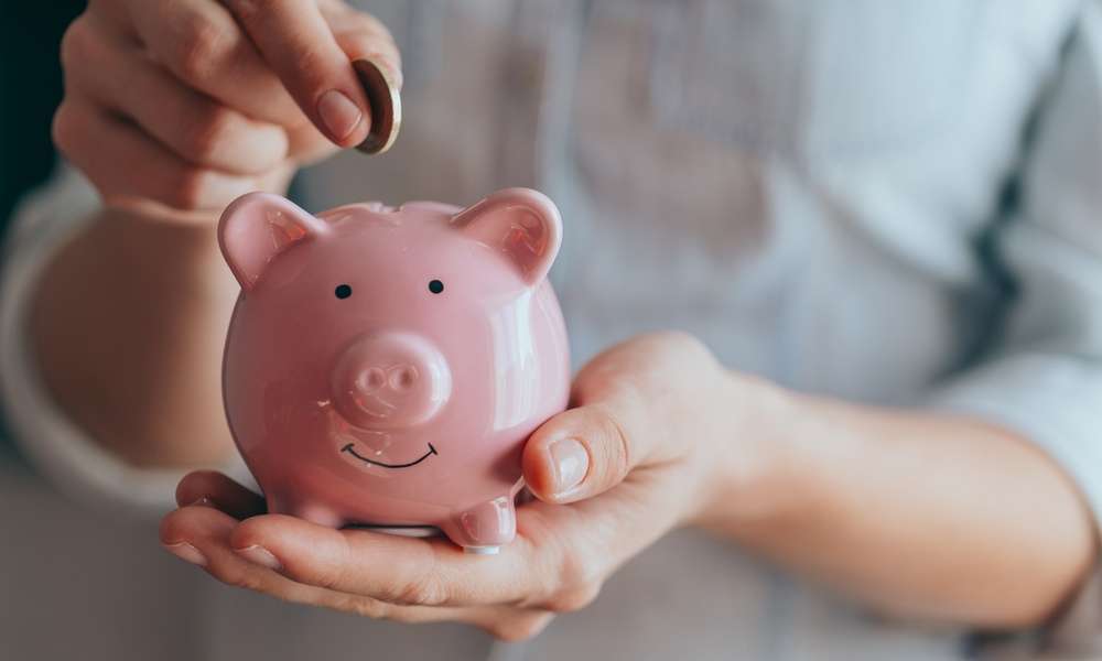 A close-up view shows a person placing a small copper coin into the slit on a small, pink piggy bank.
