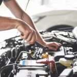 A person wearing a gray T-shirt holding a small metal wrench above the battery inside an open car hood.