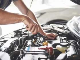 A person wearing a gray T-shirt holding a small metal wrench above the battery inside an open car hood.