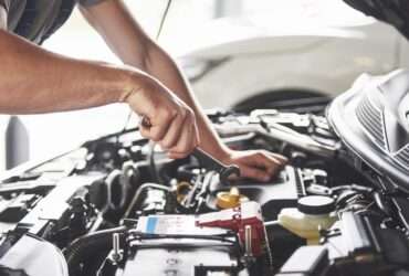 A person wearing a gray T-shirt holding a small metal wrench above the battery inside an open car hood.