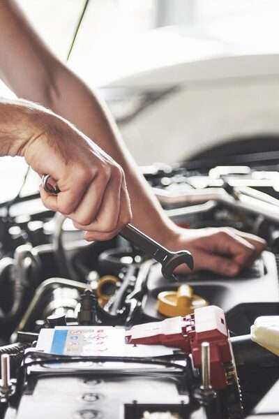 A person wearing a gray T-shirt holding a small metal wrench above the battery inside an open car hood.