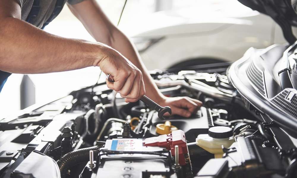 A person wearing a gray T-shirt holding a small metal wrench above the battery inside an open car hood.