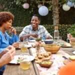 A group of adult friends or neighbors sitting around an outdoor table sharing a meal in a backyard space with trees and lights.