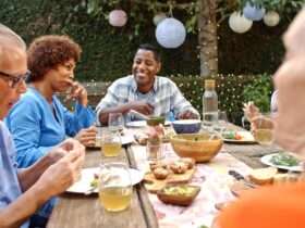 A group of adult friends or neighbors sitting around an outdoor table sharing a meal in a backyard space with trees and lights.