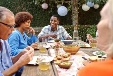 A group of adult friends or neighbors sitting around an outdoor table sharing a meal in a backyard space with trees and lights.
