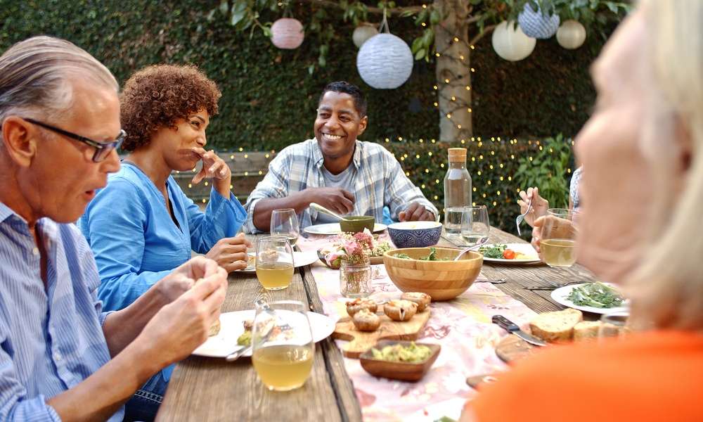A group of adult friends or neighbors sitting around an outdoor table sharing a meal in a backyard space with trees and lights.