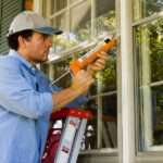 a man standing on a ladder applying caulk around an exterior window frame beside white siding and trim.
