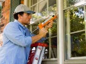 a man standing on a ladder applying caulk around an exterior window frame beside white siding and trim.