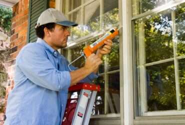 a man standing on a ladder applying caulk around an exterior window frame beside white siding and trim.