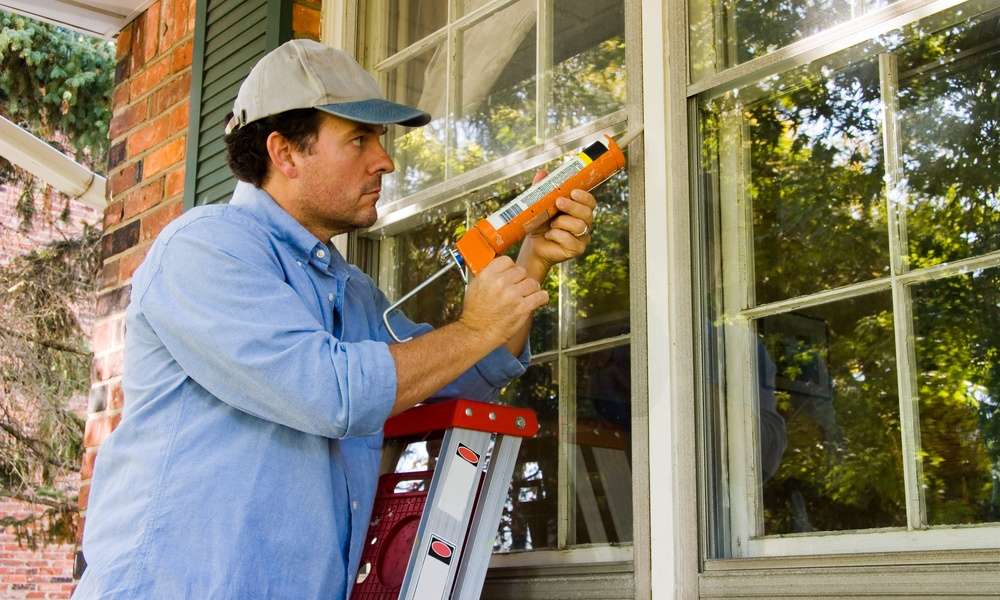 a man standing on a ladder applying caulk around an exterior window frame beside white siding and trim.