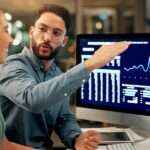 Two people sitting at a desk while one gestures toward computer monitors displaying blue financial charts.