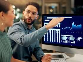 Two people sitting at a desk while one gestures toward computer monitors displaying blue financial charts.
