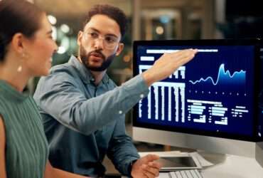 Two people sitting at a desk while one gestures toward computer monitors displaying blue financial charts.