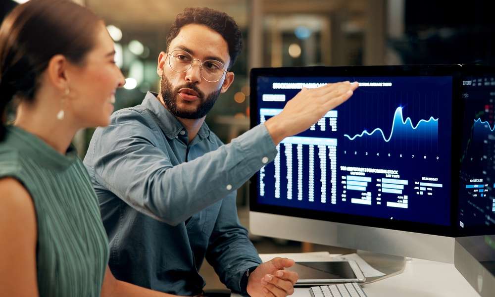 Two people sitting at a desk while one gestures toward computer monitors displaying blue financial charts.