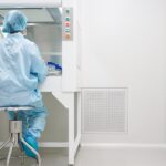 A cleanroom technician in blue protective clothing tests samples inside a laminar airflow cabinet beside a wall vent.