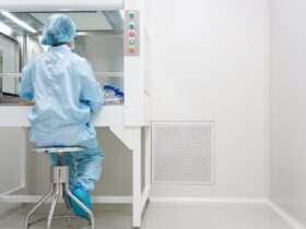 A cleanroom technician in blue protective clothing tests samples inside a laminar airflow cabinet beside a wall vent.