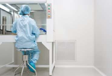 A cleanroom technician in blue protective clothing tests samples inside a laminar airflow cabinet beside a wall vent.
