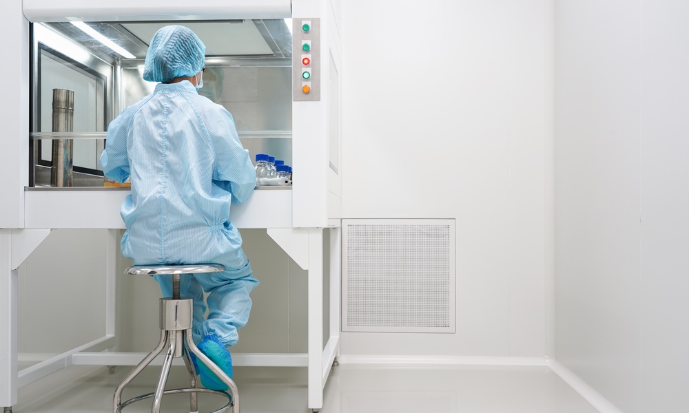 A cleanroom technician in blue protective clothing tests samples inside a laminar airflow cabinet beside a wall vent.