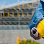 A person in a blue shirt holding a yellow hard hat and rolled papers near a blurred building under construction.