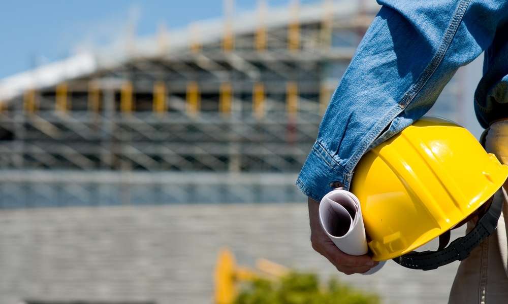 A person in a blue shirt holding a yellow hard hat and rolled papers near a blurred building under construction.