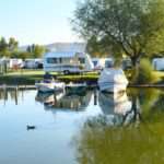 A waterfront campground lines the shore with travel trailers and boats as a large tree reflects across the calm water.