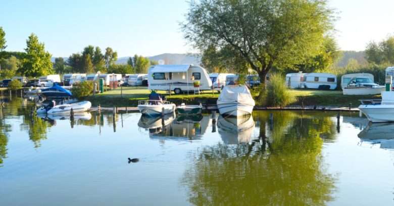 A waterfront campground lines the shore with travel trailers and boats as a large tree reflects across the calm water.