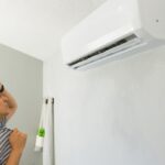 A man in a blue shirt stands indoors, holding his hand to his head, staring at an air conditioner on the wall.
