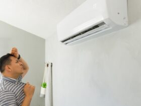 A man in a blue shirt stands indoors, holding his hand to his head, staring at an air conditioner on the wall.