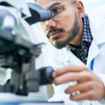 A man in glasses and a white lab coat looking into a microscope with glassware and bottles nearby in a laboratory.
