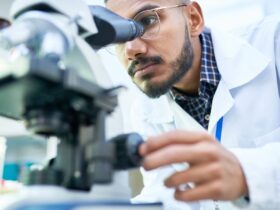 A man in glasses and a white lab coat looking into a microscope with glassware and bottles nearby in a laboratory.