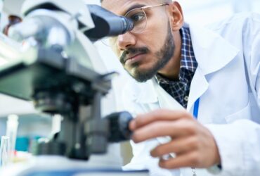 A man in glasses and a white lab coat looking into a microscope with glassware and bottles nearby in a laboratory.