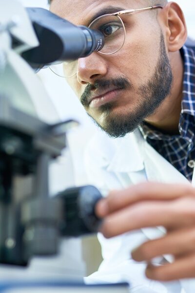 A man in glasses and a white lab coat looking into a microscope with glassware and bottles nearby in a laboratory.