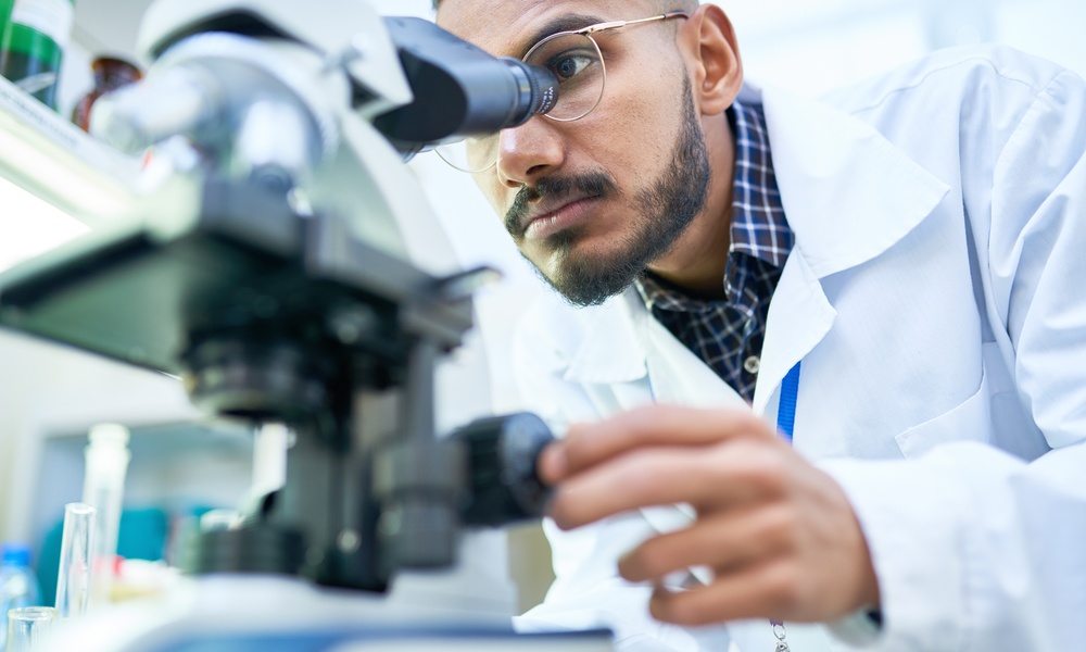 A man in glasses and a white lab coat looking into a microscope with glassware and bottles nearby in a laboratory.
