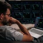 A man in glasses sitting with his laptop on his lap and a large monitor displaying green financial charts further away.