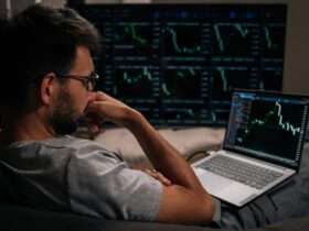 A man in glasses sitting with his laptop on his lap and a large monitor displaying green financial charts further away.