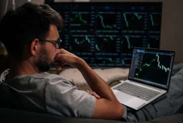 A man in glasses sitting with his laptop on his lap and a large monitor displaying green financial charts further away.