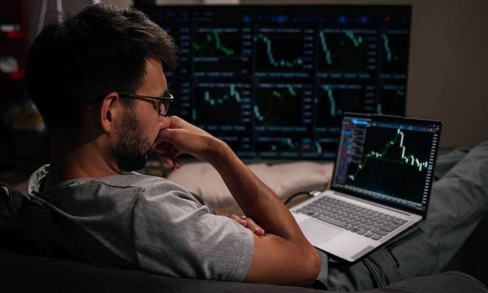 A man in glasses sitting with his laptop on his lap and a large monitor displaying green financial charts further away.